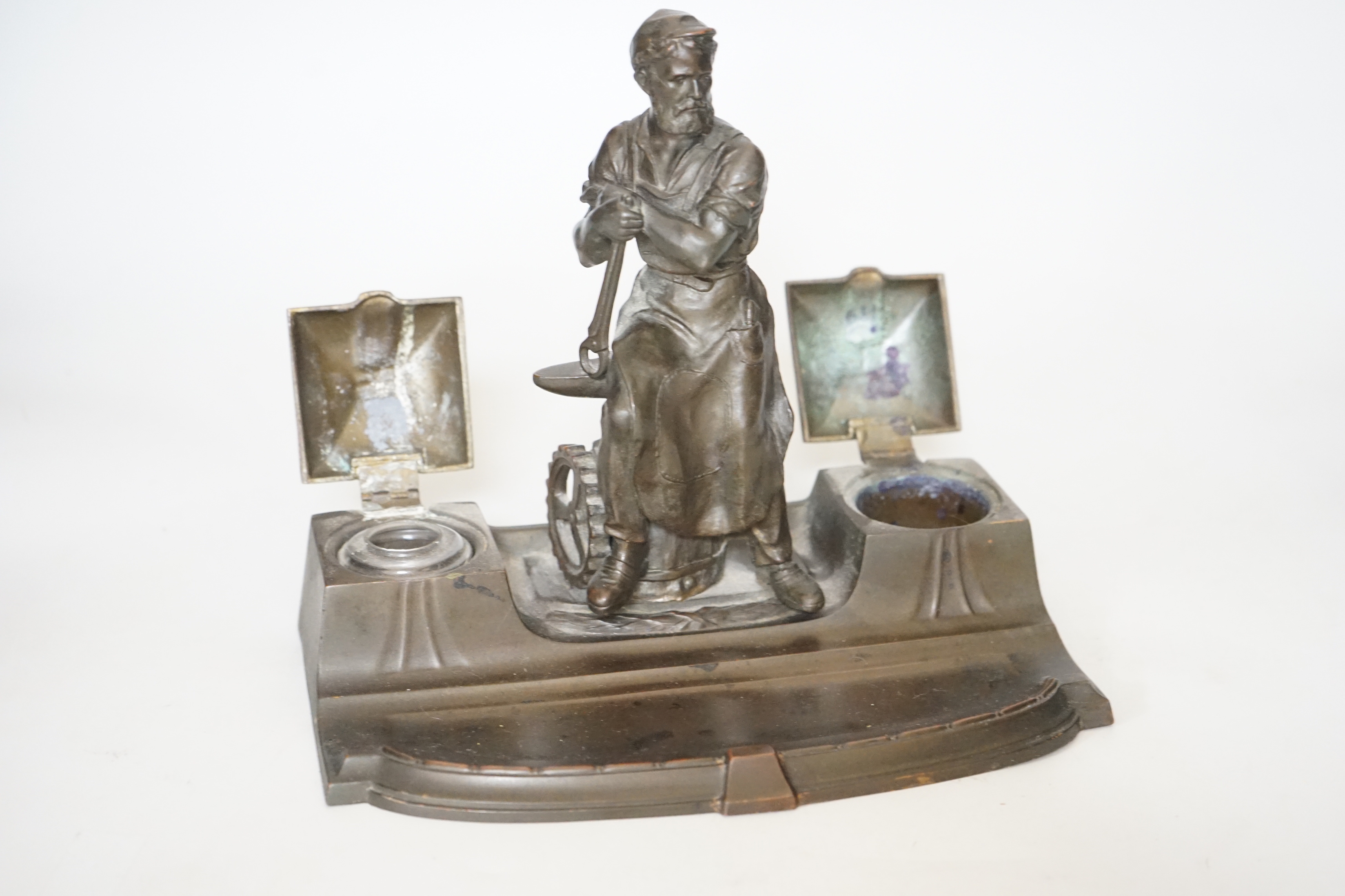 A WMF style spelter ‘Industry’ inkstand, figure with tools and an anvil, hinged ink wells either side, one glass ink bottle present, ‘1265’ cast into base, 20cm high